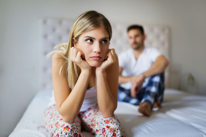 A couple sitting on a bed, the woman looking frustrated while the man sits in the background.