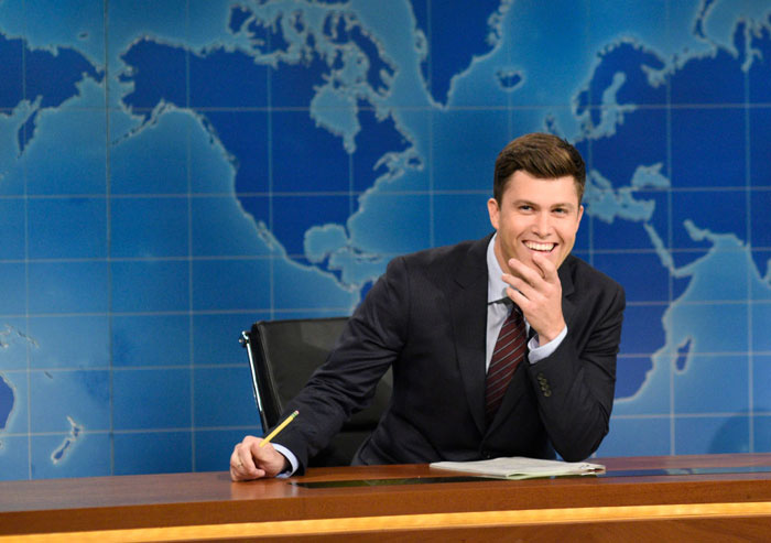 Colin Jost, Scarlett Johansson’s husband, sitting at a news desk, smiling confidently, wearing a suit and tie with a map backdrop.