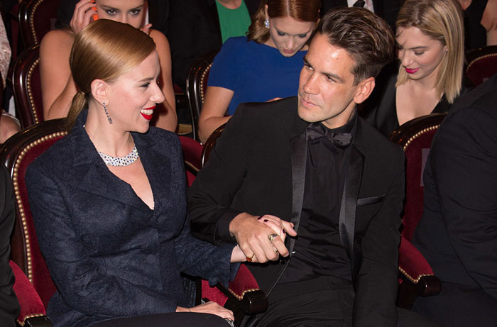 Scarlett Johansson, smiling in formal attire, holding hands with Colin Jost, her husband, dressed in a black suit, at a formal event.