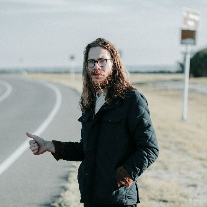 Man hitchhiking on a rural road, wearing glasses and a dark jacket, pondering the funniest joke he's heard.