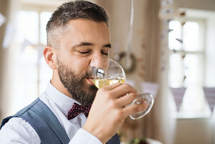 Man enjoying a glass of wine, wearing a vest and bow tie at a wedding.