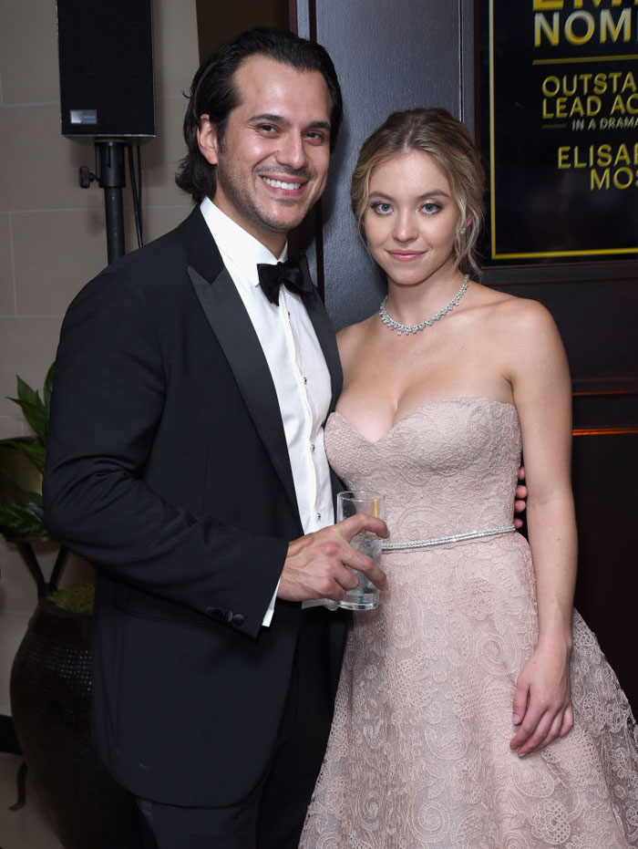 Sydney Sweeney, wearing a strapless light pink lace gown with a delicate necklace, poses beside Jonathan Davino, dressed in a black tuxedo and bow tie. They smile together at an event.