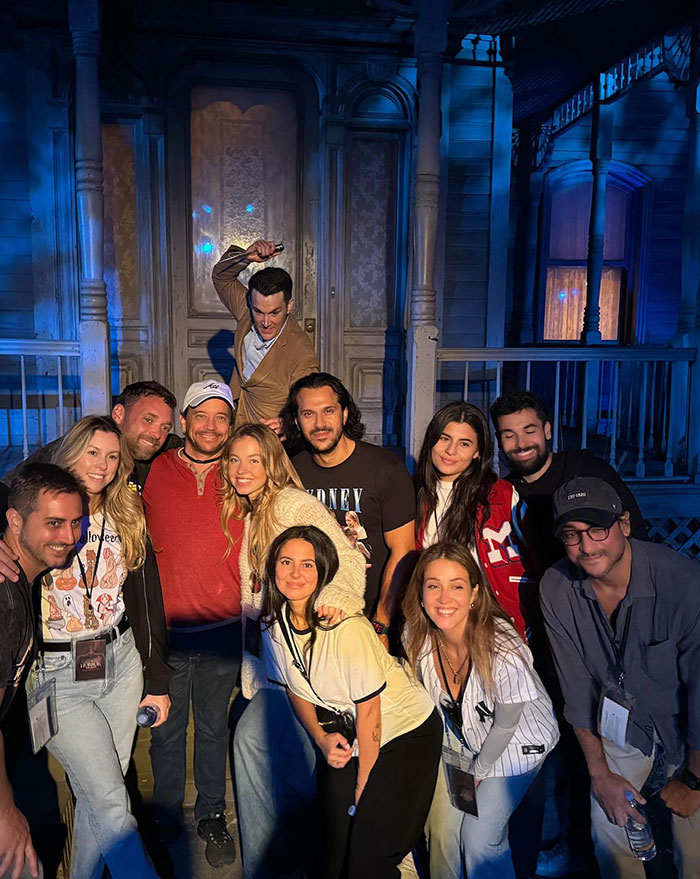  Sydney Sweeney and Jonathan Davino pose with a group of friends in front of a spooky, dimly lit house set, likely part of a haunted attraction. The group, smiling and gathered closely, is accompanied by an actor in costume behind them, adding to the eerie ambiance. Everyone appears excited and engaged, dressed casually, and some wear themed attire, adding to the fun, immersive experience.