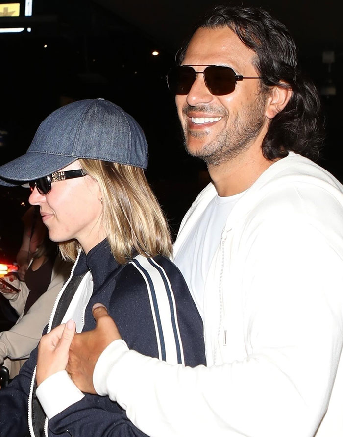 Sydney Sweeney, dressed casually in a dark jacket with white stripes and a denim baseball cap, is seen walking closely with Jonathan Davino, who wears a white hoodie and sunglasses. Jonathan smiles broadly as he wraps an arm affectionately around Sydney's shoulders. The setting appears to be at night, with both dressed in relaxed attire, enjoying their time together.