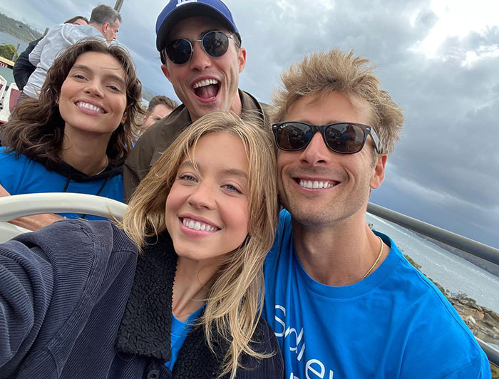  Sydney Sweeney poses with friends outdoors, all smiling and looking cheerful. Sydney is dressed in a dark jacket over a blue shirt, while her friends wear blue shirts and sunglasses, with one in a cap. They appear to be enjoying a casual day together with an ocean view in the background, under a cloudy sky.