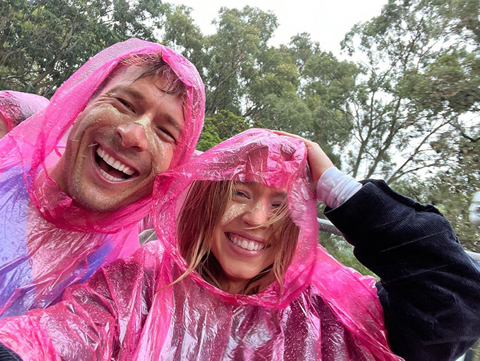  Sydney Sweeney and a friend are wearing bright pink rain ponchos, laughing together in an outdoor setting. Both have sand on their faces, suggesting they recently had a playful or windy experience at the beach. They look happy and carefree, enjoying the rainy day surrounded by trees.