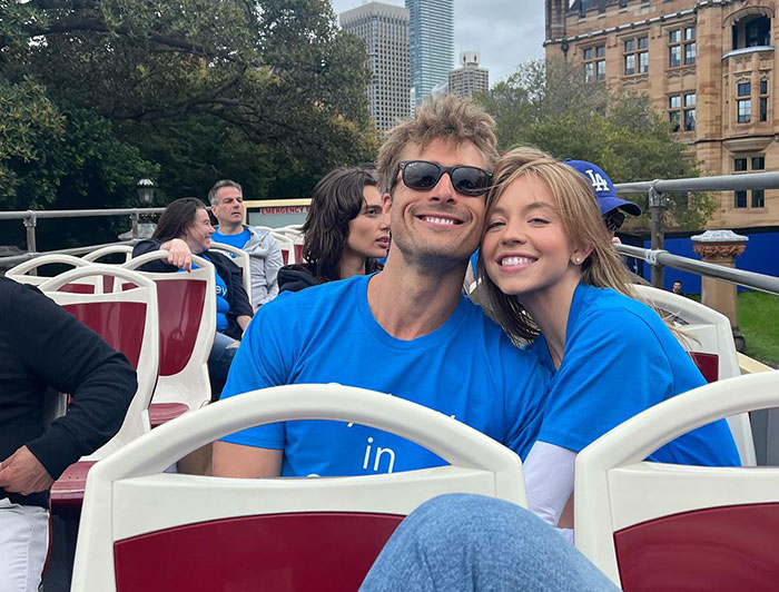 Sydney Sweeney and a friend sit together on an open-top tour bus, both wearing matching blue shirts. Sydney leans affectionately on her friend, smiling broadly, while he wears sunglasses and grins. The setting appears to be in a city, with skyscrapers and historic buildings visible in the background. Other passengers can be seen enjoying the scenic ride.