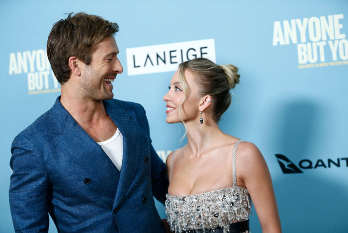 Sydney Sweeney, in a sparkling gown, smiles and gazes at her co-star, who is dressed in a blue suit, at the Anyone But You event. They pose together against a light blue backdrop with sponsor logos.