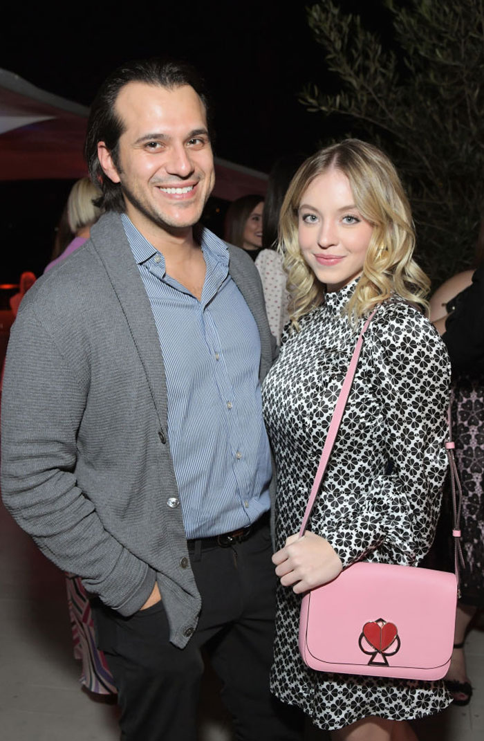 Sydney Sweeney, dressed in a black-and-white patterned long-sleeve dress and carrying a pink bag with a heart design, stands beside Jonathan Davino, who is wearing a blue striped shirt with a gray cardigan. They smile together at an event.