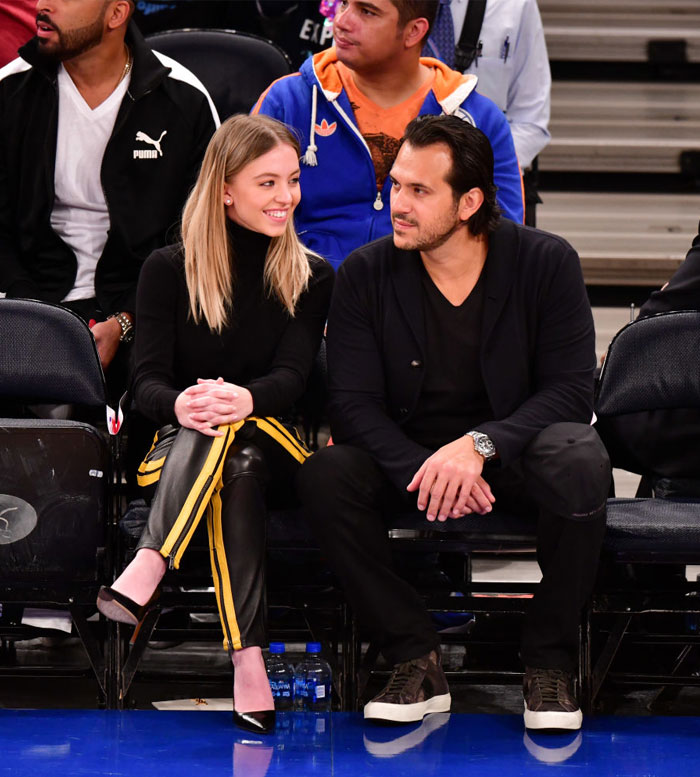 Sydney Sweeney, dressed in a black turtleneck and black pants with yellow side stripes, sits courtside at a sports event, smiling at Jonathan Davino, who is dressed in black attire with a watch on his wrist. They appear engaged in conversation amidst the crowd.