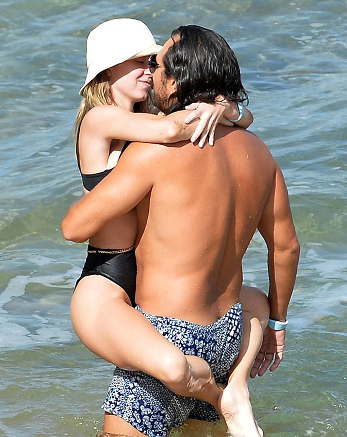  Sydney Sweeney, wearing a white sun hat and a black swimsuit, is held in the arms of Jonathan Davino, who is shirtless and wearing patterned swim trunks. They embrace affectionately in the water, smiling at each other.