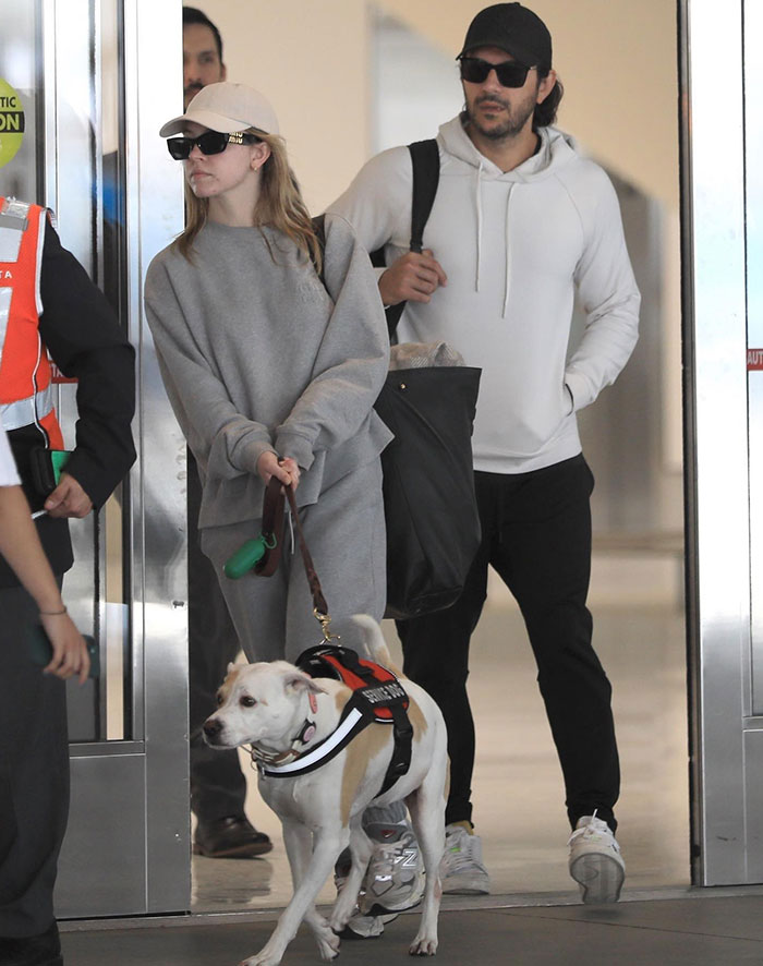 Sydney Sweeney, dressed in a gray sweatshirt and pants with a beige baseball cap and sunglasses, holds the leash of a white dog wearing a harness. Beside her, Jonathan Davino is dressed in a white hoodie, black pants, and a black cap, carrying a large bag over his shoulder. They appear to be at an airport or public indoor space, walking together with their dog.