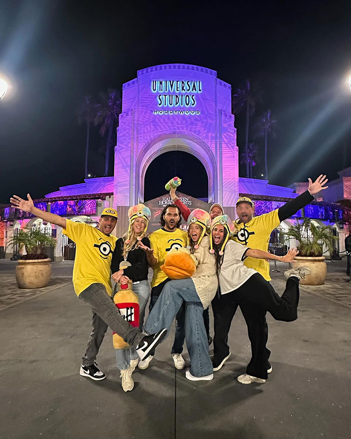 A group of people, including Sydney Sweeney and Jonathan Davino, pose excitedly in front of the illuminated Universal Studios Hollywood entrance at night. They are dressed in yellow Minion-themed attire, some wearing Minion hats and others in Minion shirts, showing playful expressions and energetic poses, celebrating their visit to the theme park.