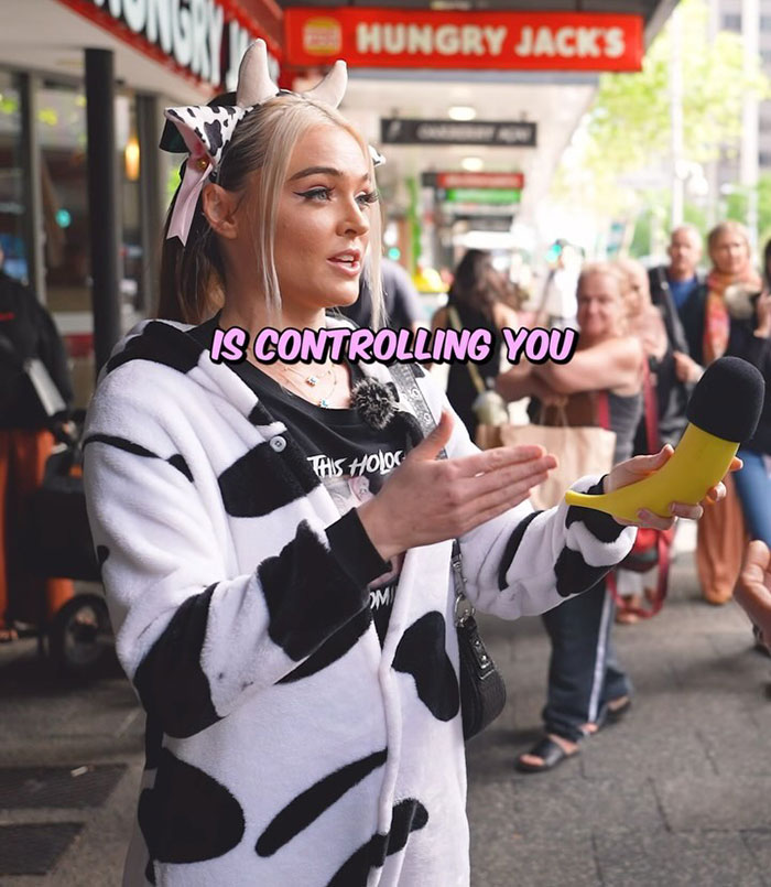 Vegan activist in cow-patterned outfit holding a microphone, advocating against animal slavery on a busy street.