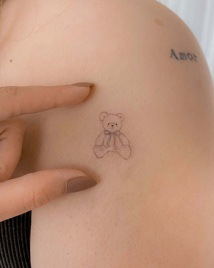 Small teddy bear tattoo on woman's shoulder next to the word "Amor," showcasing subtle tattoo ideas for women.