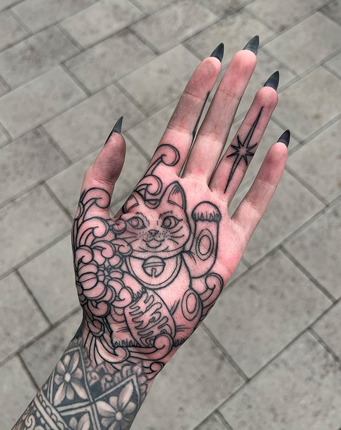 A tattoo idea for women with a hand tattoo featuring a black ink design of a lucky cat and patterns.