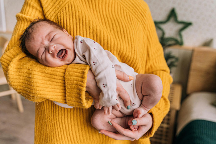 Baby held by mother-in-law in a yellow sweater, appearing upset while being cradled in a cozy room.