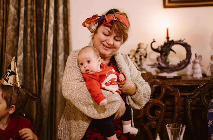 Grandmother holding baby in a red outfit during holiday gathering, smiling and looking affectionate.