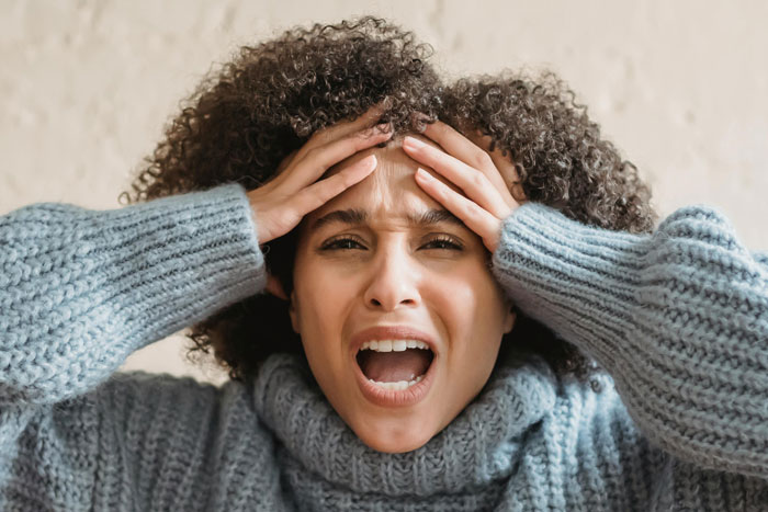 Frustrated mom with hands on head, wearing gray sweater, expressing stress.