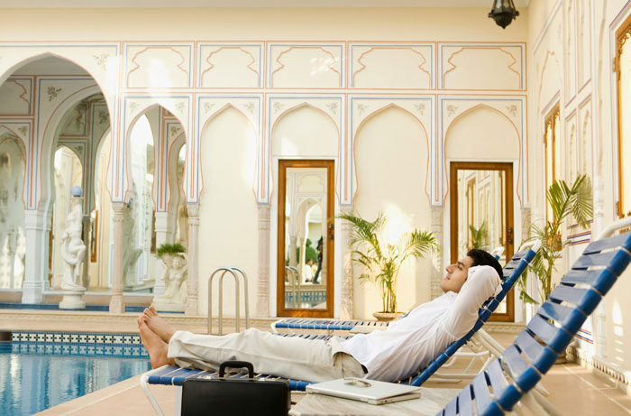 Man relaxing by a luxury hotel pool, lounge chair and laptop beside him, enjoying a fancy vacation.