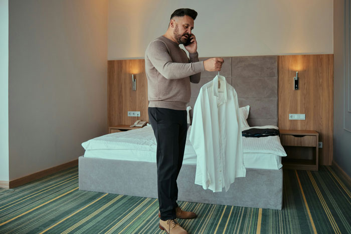 Man in hotel room on phone, holding shirt, during vacation disrupted by unexpected family visit.