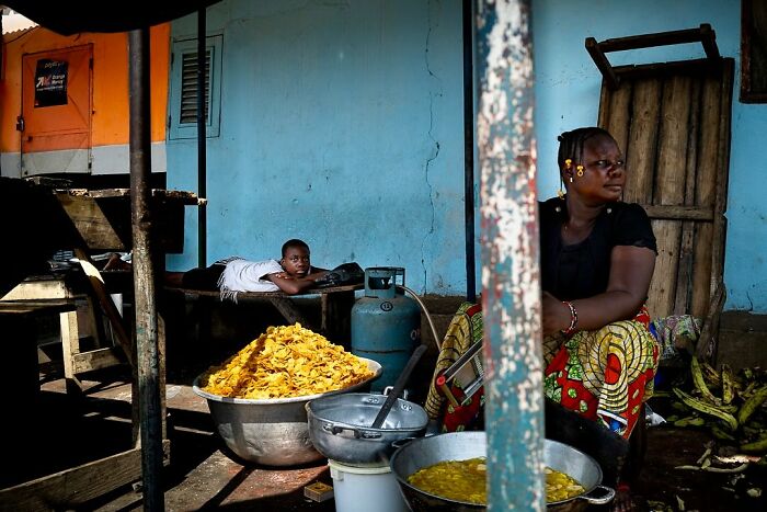 A woman sits beside bowls of sliced plantains in a vibrant setting, with a child lying nearby, captured by Ivan Margot.