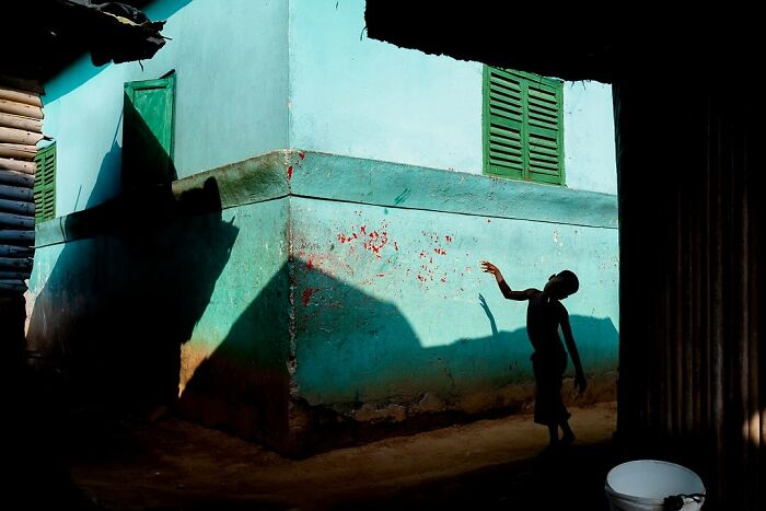 Silhouette of a person reaching towards a vibrant teal wall with green shutters, casting a shadow, by Ivan Margot.