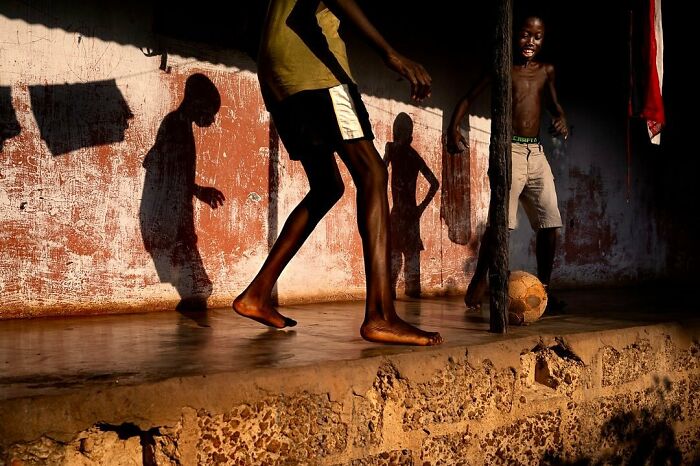 Children playing soccer, casting shadows against a weathered wall in a vibrant photograph by Ivan Margot.