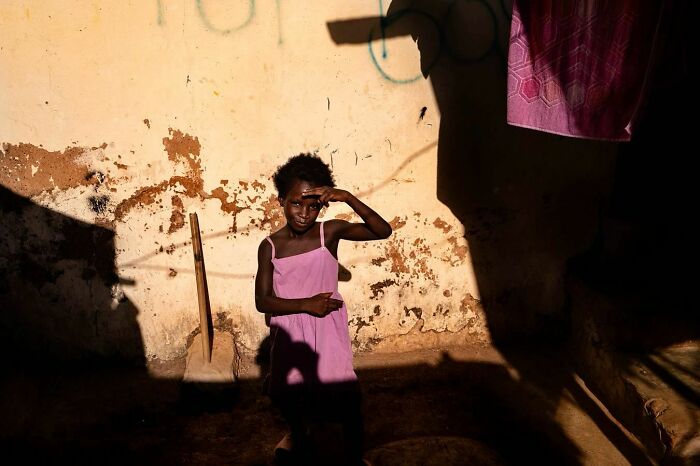 Child in a pink dress shielding eyes from sunlight, captured in vibrant photograph by Ivan Margot.