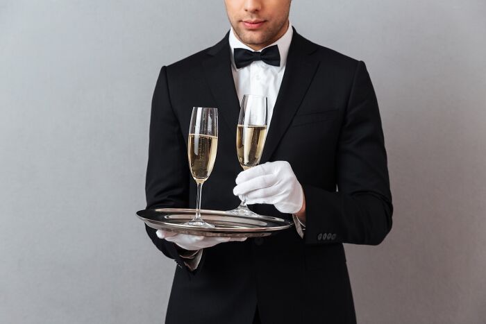 Butler in tuxedo holding a tray with champagne, indicating wealth.