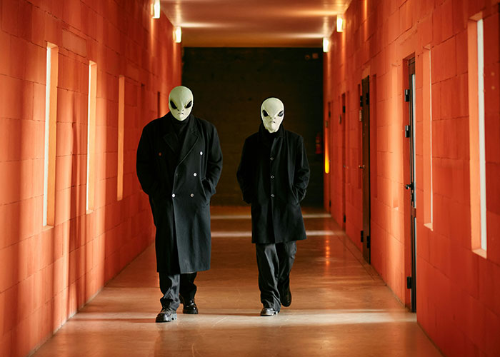 Two figures in alien masks and black coats walking down a mysterious hallway.