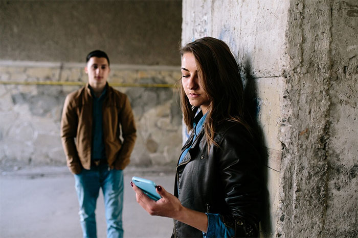 Woman leans against a wall, looking at her phone, while a man stands in the background.