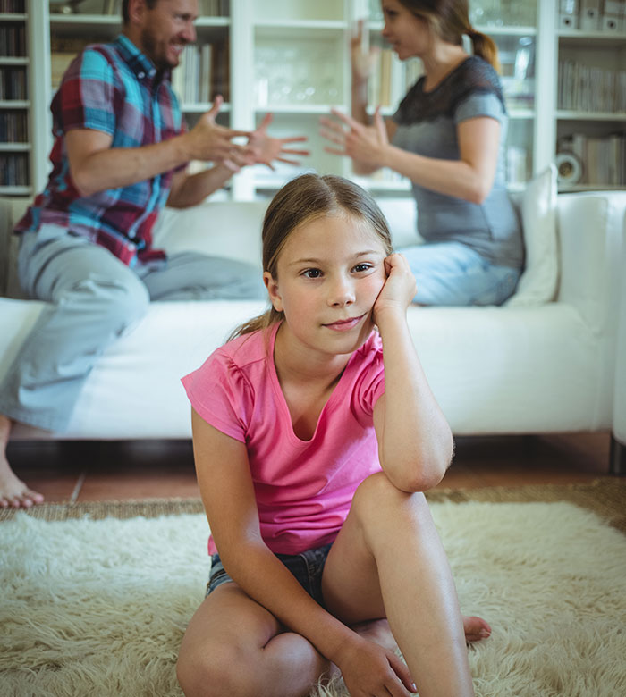 Child sitting on rug with adults arguing in the background, highlighting family tension and dynamics.