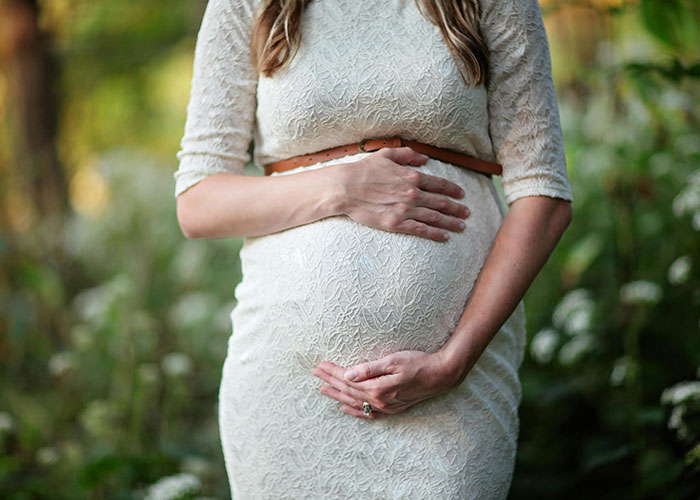 Pregnant woman in lace dress standing in garden, hands on her belly, serene and thoughtful.