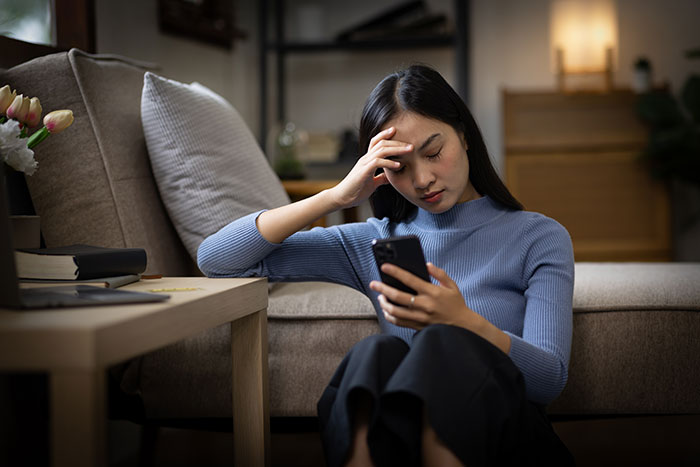 A concerned woman in a blue sweater, seated on the floor and looking at her phone in an apartment.