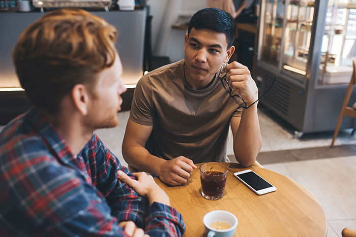 Two men at a café table, one holding glasses and listening, illustrating communication and cultural judgment.