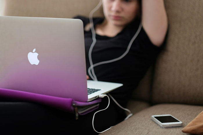 Gen A individual with laptop and headphones, focused on screen, phone lying on sofa beside them.