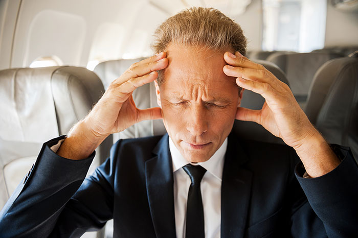 Frustrated passenger in a suit with hands on temples, seated on a plane.