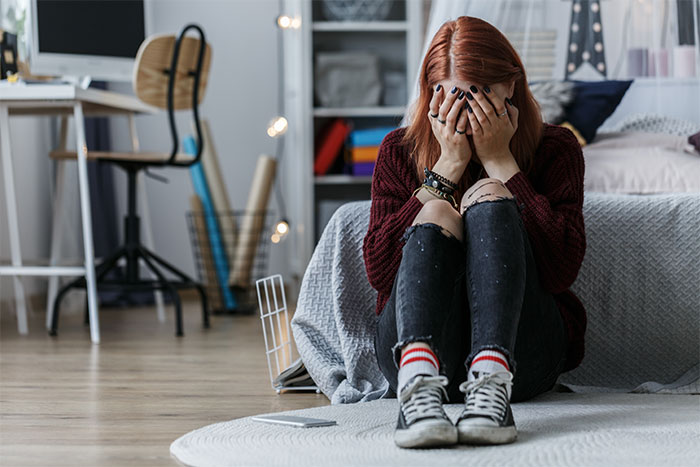 Teen sitting on bedroom floor, looking distressed, hiding face with hands.