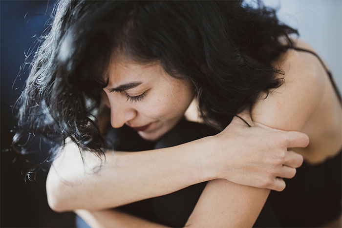 A woman with dark hair sits curled up, appearing distressed, in relation to hidden birth control pills.