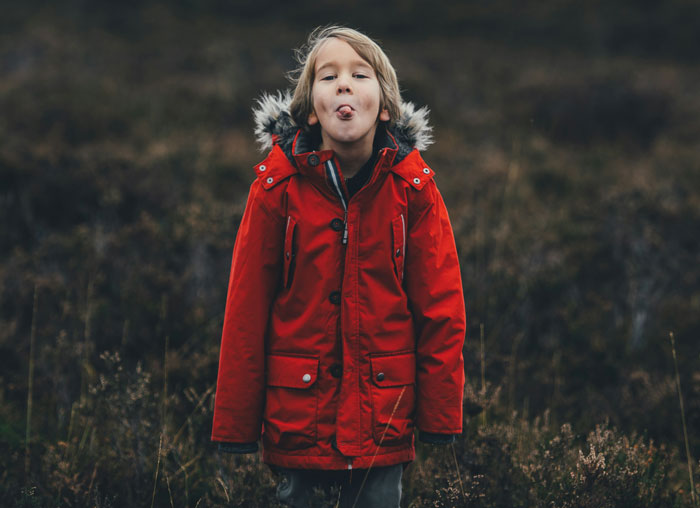 Child from Gen A in a red coat sticking tongue out playfully outdoors.