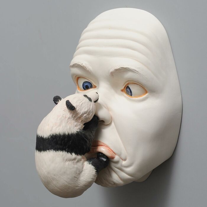 Sculpture blending realism and surrealism: panda touching a human face on a gray wall.