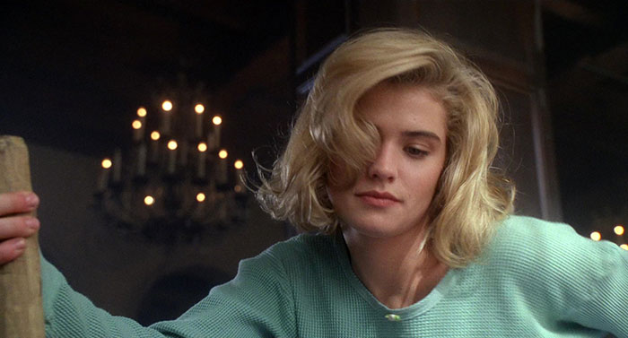 A woman with short blonde hair wearing a green sweater, embodying a strong female protagonist in a dimly lit room.