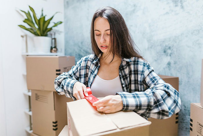 Woman sealing a cardboard box with red tape, standing in a room with several moving boxes and a plant.