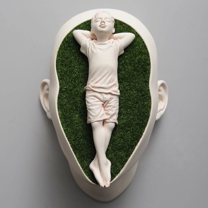 Realistic and surreal sculpted head with a relaxed child figure on grassy surface.