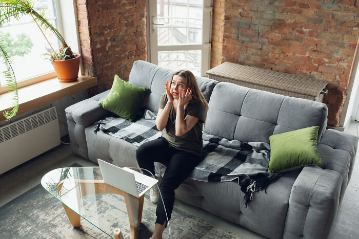 Woman on gray sofa, looking surprised, hands on cheeks, experiencing a terrible Christmas moment at home.