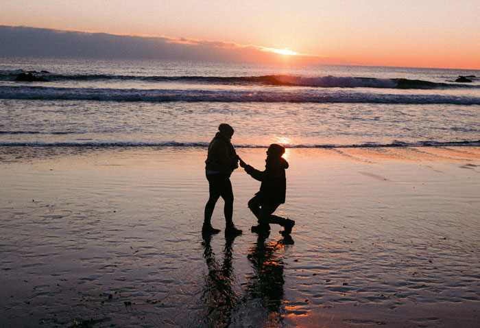 Sunset beach proposal with silhouetted couple, ocean waves, and golden sky, capturing genuine emotions and surprises.