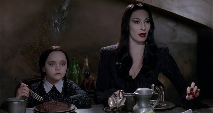 A strong female protagonist with a young girl at a dining table in a dimly lit room, conveying a gothic family dynamic.