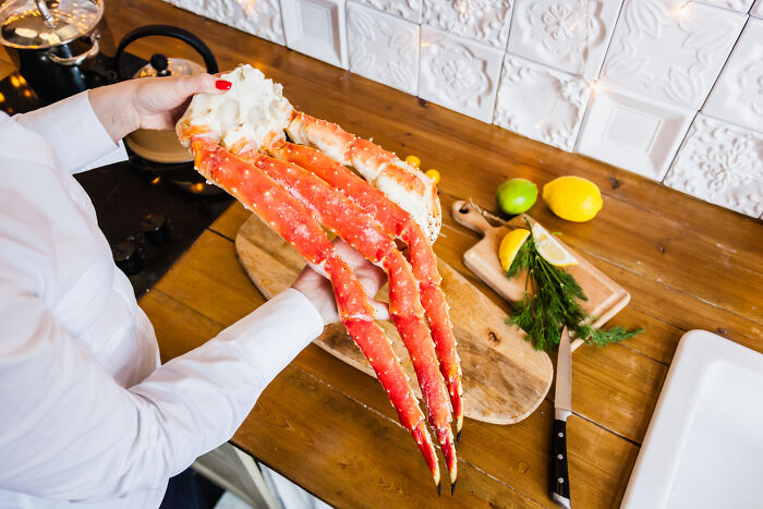 Woman holding large crab legs in a kitchen with lemons and herbs, pondering cooking ideas.