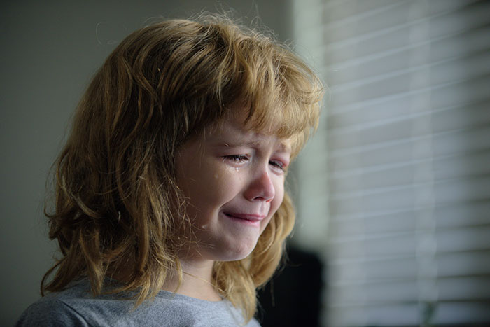 Child crying near window blinds, expressing sadness, related to affair child and Christmas gift context.